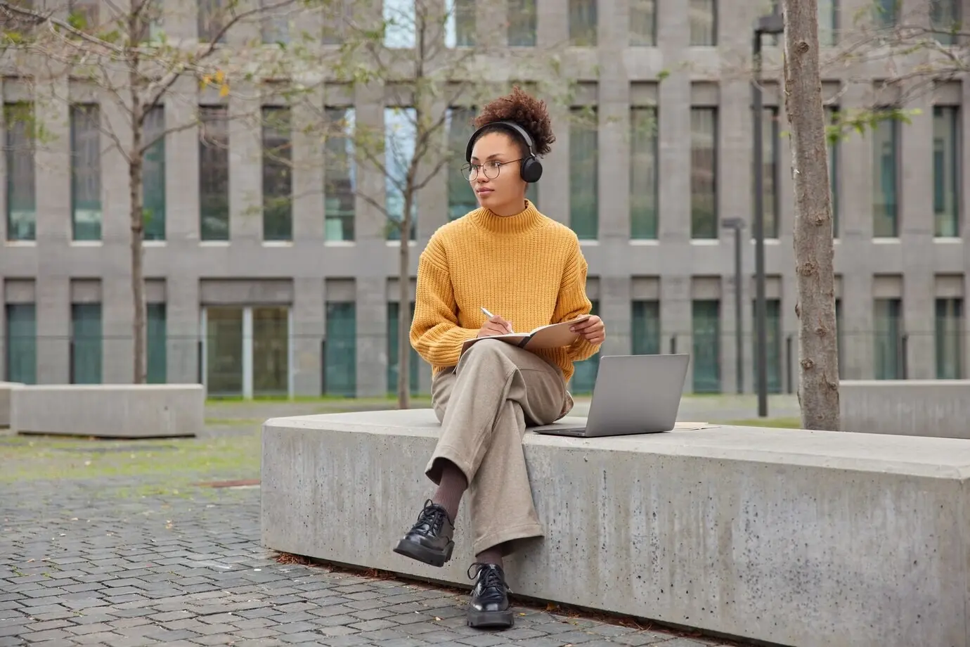 Schülerin schreibt Notizen in ein Notizbuch, hört ein Webinar, bereitet sich auf eine Prüfung vor, posiert im Freien, arbeitet am Laptop, posiert vor urbaner Kulisse, plant den Organisationsprozess.